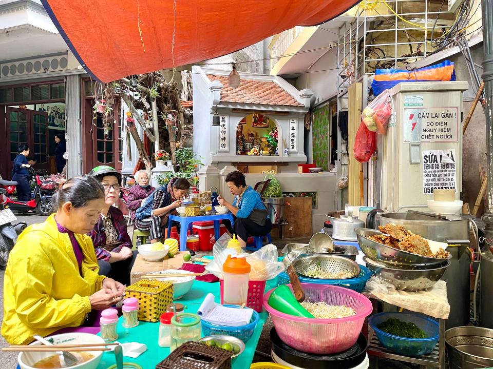 Women eating on stools outside  Bún Cá Sâm Cây Si street kitchen, next to shrine, in Hanoi