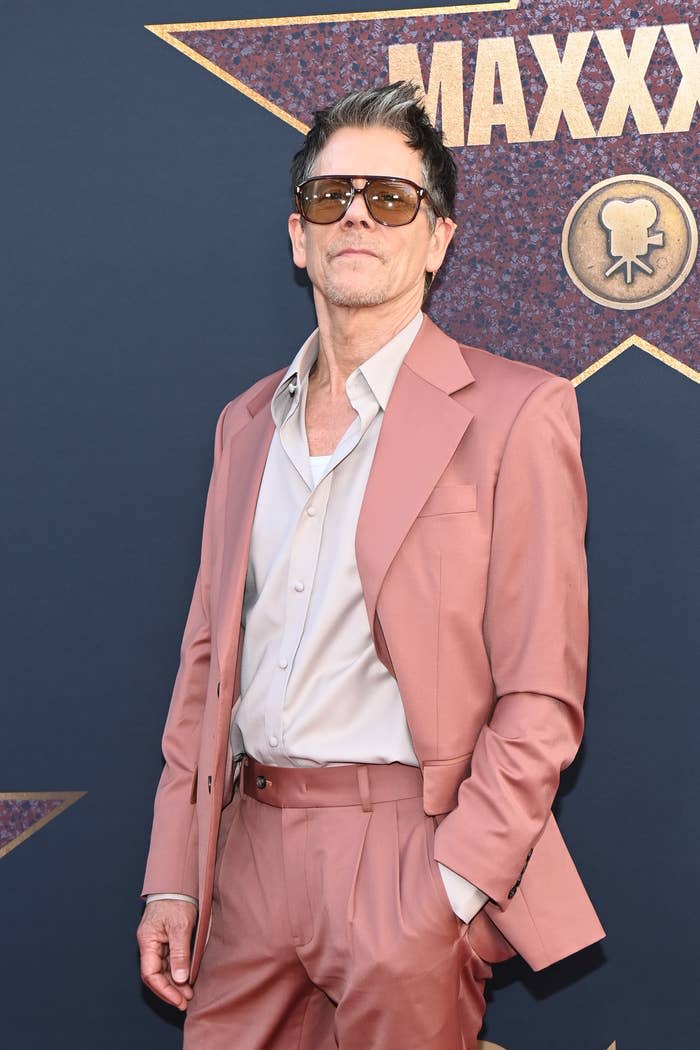 Kevin Bacon stands on the red carpet in a stylish suit with sunglasses, posing confidently