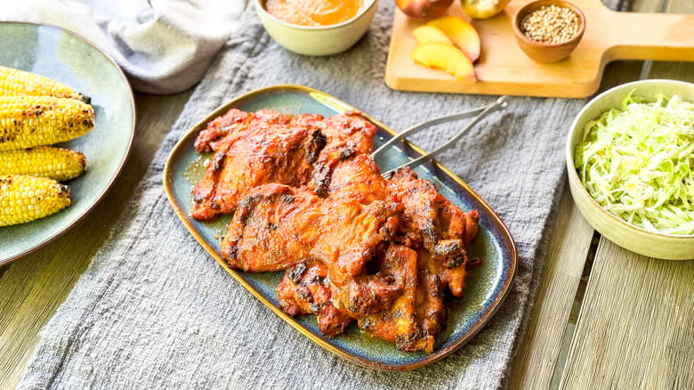 Peach bbq chicken thighs on platter with grilled corn, coleslaw, and ingredients on table