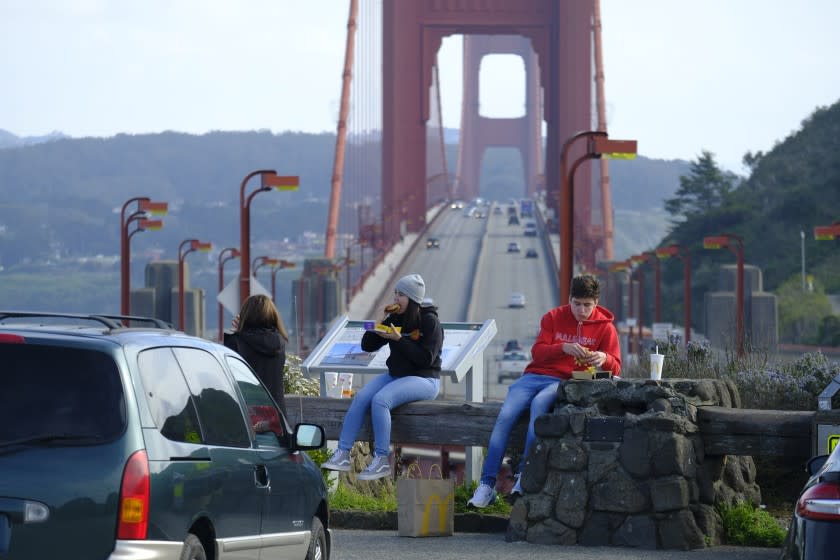 People eat while sitting at a vista point by the Golden Gate Bridge Friday, March 27, 2020, in Sausalito, Calif. The surge of coronavirus cases in California that health officials have warned was coming has arrived and will worsen, Gov. Gavin Newsom said Friday, while the mayor of Los Angeles warned that by early next week his city could see the kind of crush that has crippled New York.(AP Photo/Eric Risberg)