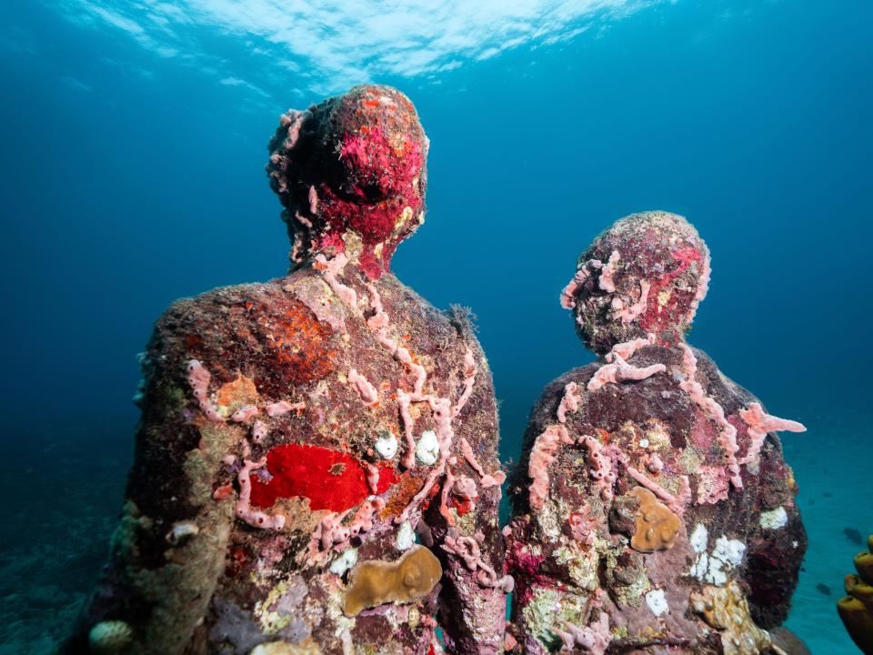 Two colorful, human-like underwater sculptures covered in coral and other marine life.