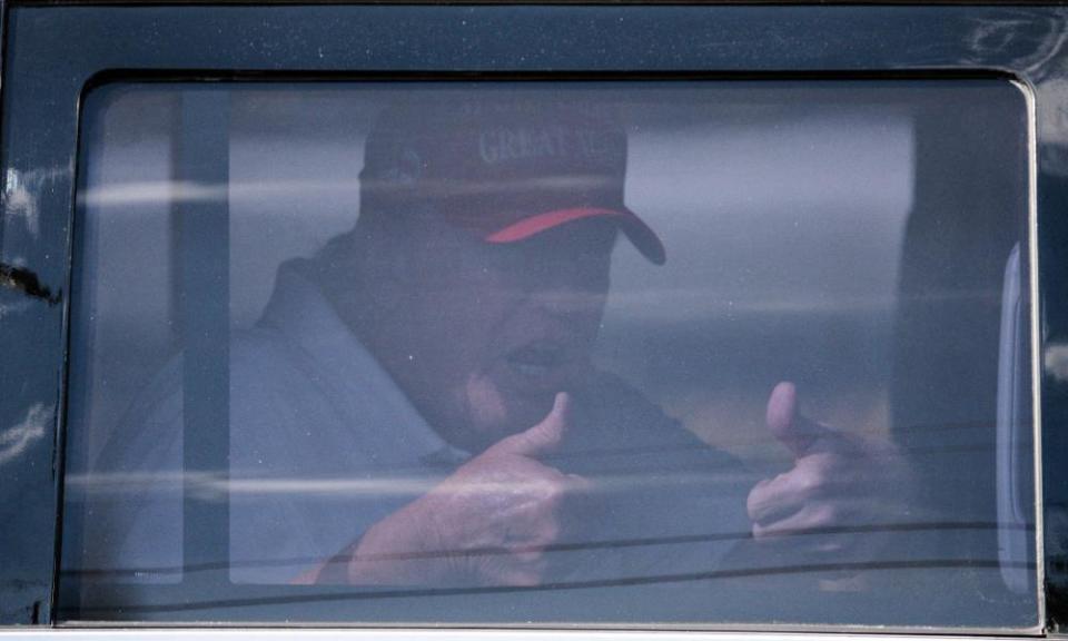 Trump in a red make America great again hat giving two thumbs up from inside his limousine