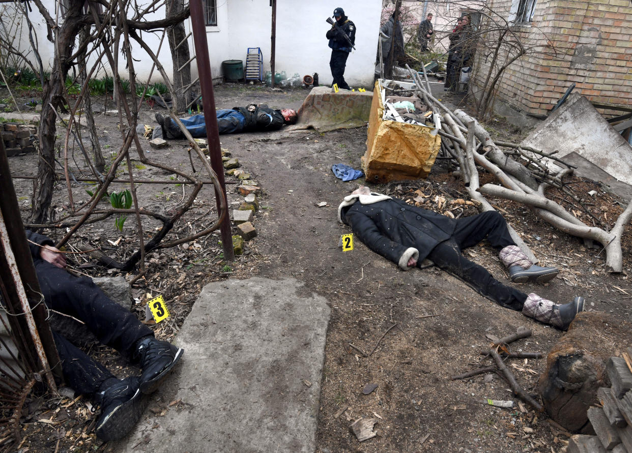 The bodies of two men lie near a damaged home in Bucha as a soldier stands nearby. 