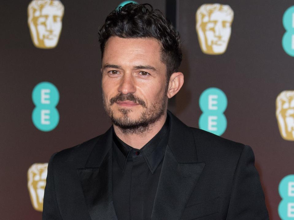Orlando Bloom wearing an all-black suit in front of a brownish and blue background