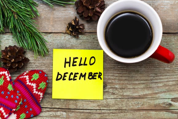 Post-it note that says Hello December on wood surface, surrounded by coffee cup, glove, and pinecones.