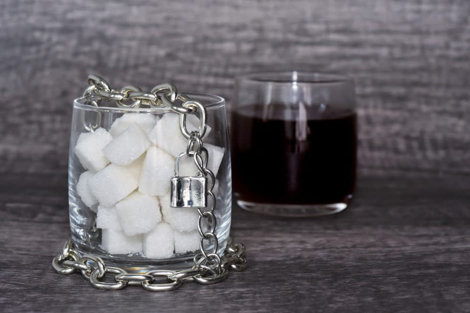 Ein mit Zuckerwürfeln gefülltes Glas mit Kette und Vorhängeschloss, dahinter ein Glas mit Kaffee