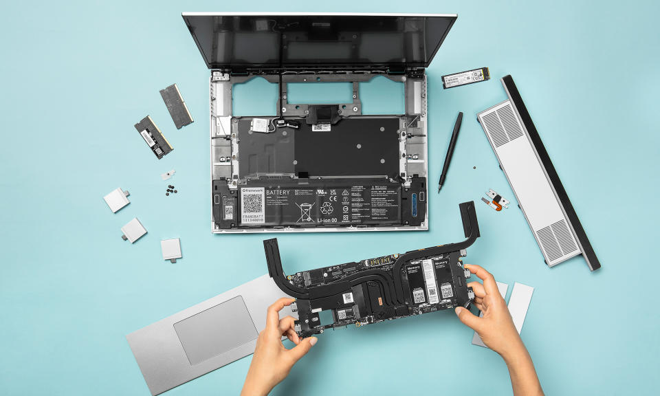 Marketing photo for the Framework Laptop 16, showing a top-down view of a person swapping out its parts. The laptop is partially dismantled with pieces all around. Two hands emerging from bottom of frame hold a module, including memory and graphics.