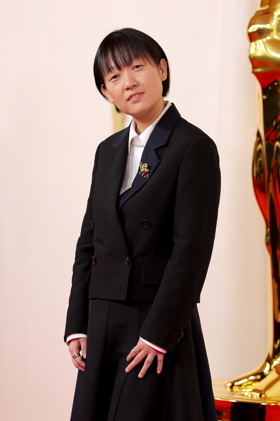 A woman stands on the red carpet in a formal black suit with a white shirt, next to a golden statue resembling an Oscar