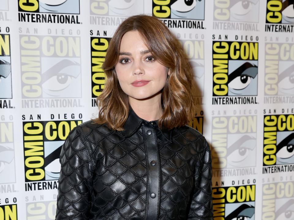 jenna coleman on the san diego comic-con red carpet, wearing a black leather dress hat buttons up to her neckline with a collar. her hair is shoulder length and she's smiling slightly at the camera