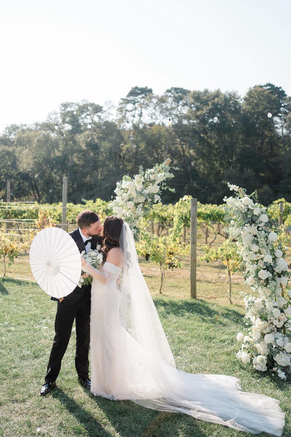 A bride and groom kiss in front of a floral arch. They hold a parasol in front of them.