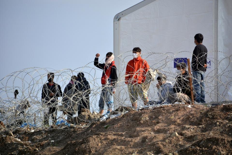 Migrants stand behind a fence at Karatepe refugee camp, on the northeastern Aegean island of Lesbos, Greece, Monday, March 29, 2021. The European Union's home affairs commissioner is visiting asylum-seeker facilities on the eastern Greek islands of Samos and Lesbos amid continuing accusations against Greece of illegal summary deportations. (AP Photo/Panagiotis Balaskas)