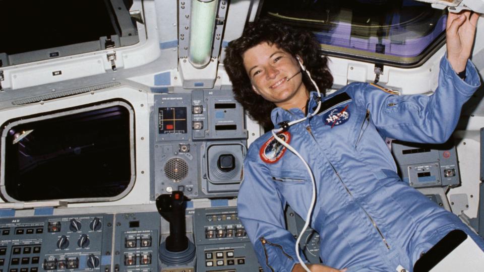 sally ride smiling in the space shuttle cockpit alongside controls, a joystick and a couple of windows