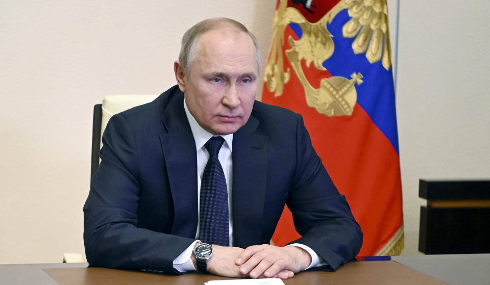 President Vladimir Putin, looking emotionless, sits, hands clasped, before a heraldic Russian flag.