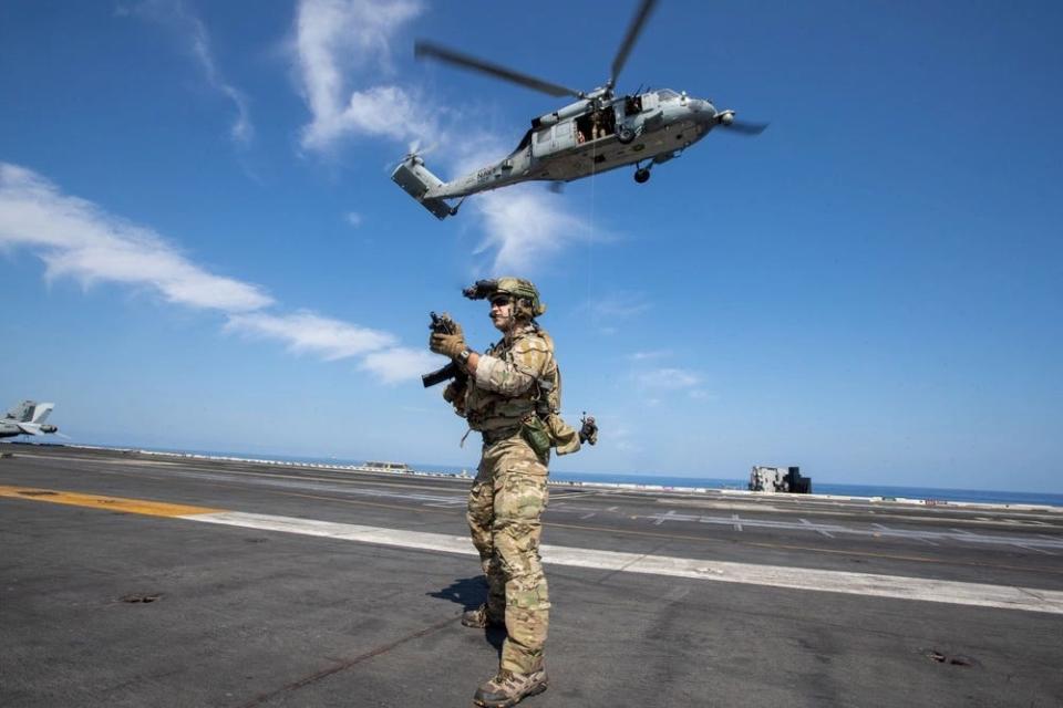 a sailor in uniform stands on the flight deck of a military aircraft carrier while a helicopter in the background hoists another sailor
