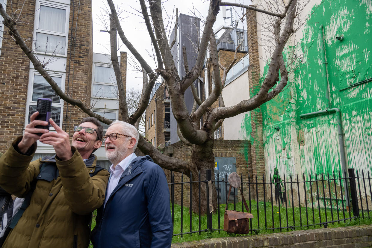 LONDON, UNITED KINGDOM - March 18: Former Labour Party politician Jeremy Corbyn (R) poses next to a new tree mural artwork appeared on a building that has been confirmed to be the work of the artist Banksy at Finsbury Park in London, United Kingdom on March 18, 2024. The artwork has been painted in front of a cut down tree to look like green foliage also matching the green used by Islington Council for signs. (Photo by Stringer/Anadolu via Getty Images)