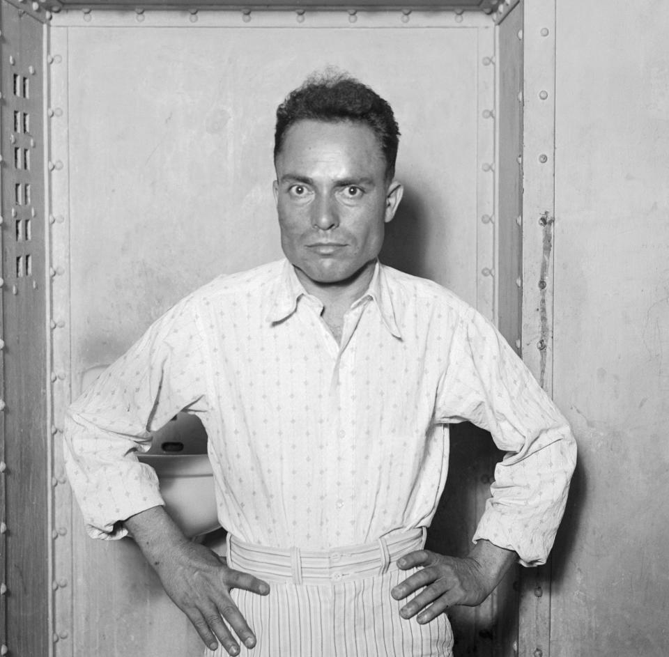 Guiseppe Zangara with his hands on his hips, wearing a collared shirt and striped pants. The background shows what appears to be a metal door