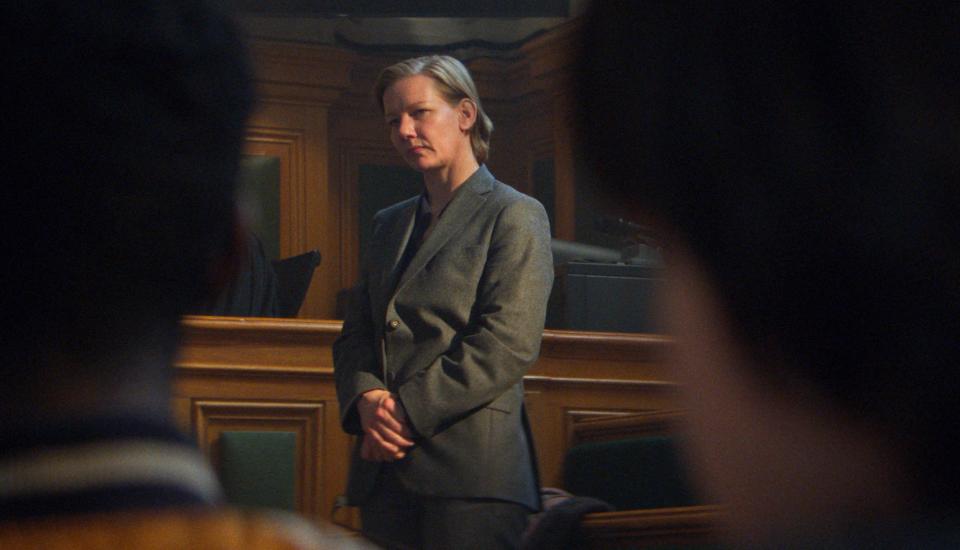 Still Sandra Hüller in "Anatomy of a Fall" standing in a court box, framed by people watching her in the gallery of a courtroom.