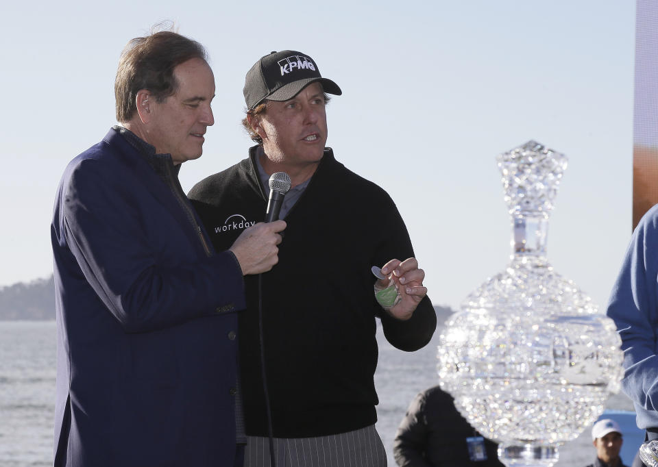 Phil Mickelson holds up a silver dollar that belonged to his grandfather during an awards ceremony on the 18th green of the Pebble Beach Golf Links after winning the AT&T Pebble Beach Pro-Am golf tournament Monday, Feb. 11, 2019, in Pebble Beach, Calif. At left is broadcaster Jim Nantz. (AP Photo/Eric Risberg)