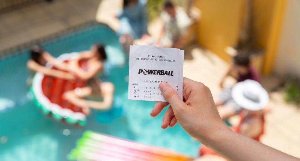 Hand holds winning Powerball ticket in front of pool with friends.