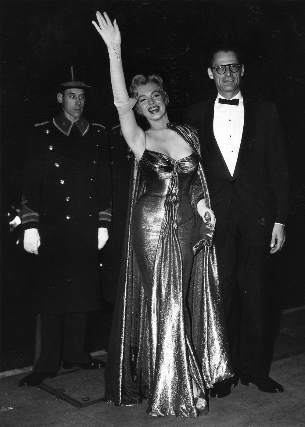 Marilyn Monroe wearing a gold lame gown and waving next to Arthur Miller.