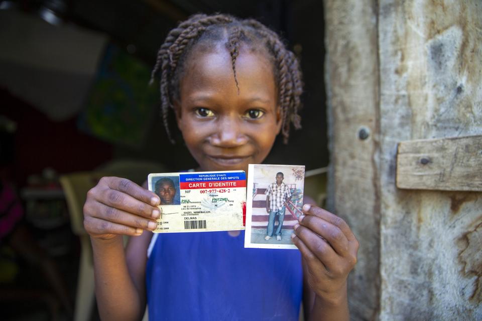 Eight-year-old Naffetalie Paul holds up an ID and photo of her late father Fritznel Paul, who died of cholera at age 39 when she was a newborn, as she stands in the doorway of her aunt's home where her father supported his two sisters and their six children in Mirebalais, Haiti, Monday, Oct. 19, 2020. Ten years after a cholera epidemic swept through Haiti and killed thousands, families of victims still struggle financially and await compensation from the United Nations as many continue to drink from and bathe in a river that became ground zero for the waterborne disease. (AP Photo/Dieu Nalio Chery)