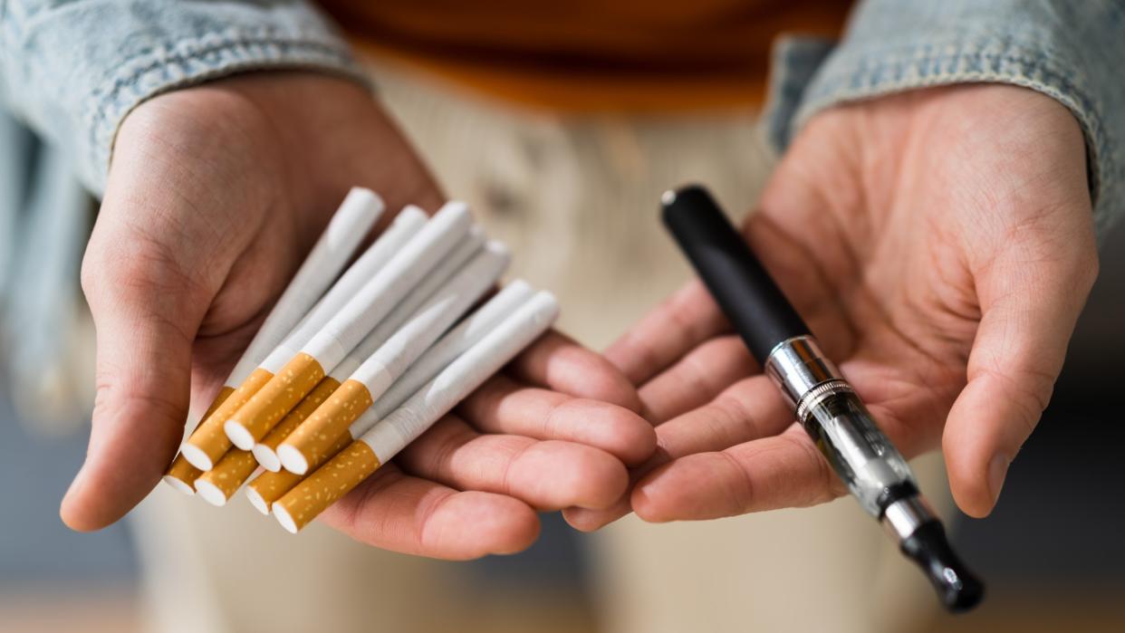  Close up of a pair of hands holding smoking parafernalia. In the left hand is a vape pen and in the right hand there is a handful of cigarettes. 