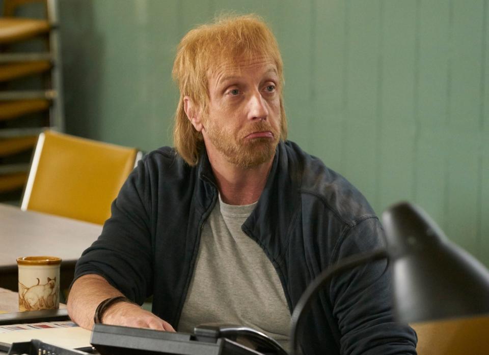 Man sitting at a desk with phones, looking pensive in a TV show scene