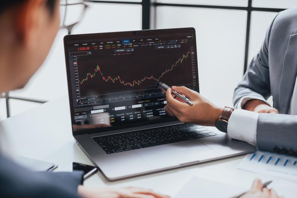 A financial advisor uses a pen to show a client the low point of a price chart displayed on a laptop. 