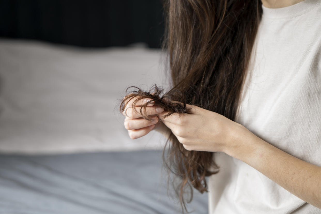 A woman appears concerned as she closely inspects the split ends of her long, wavy brunette hair, possibly considering remedies for hair damage.