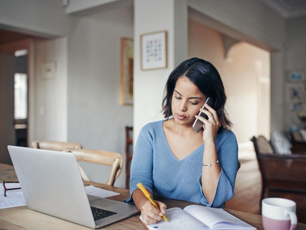 Shot of a young woman using a mobile phone, laptop and writing notes while working from home