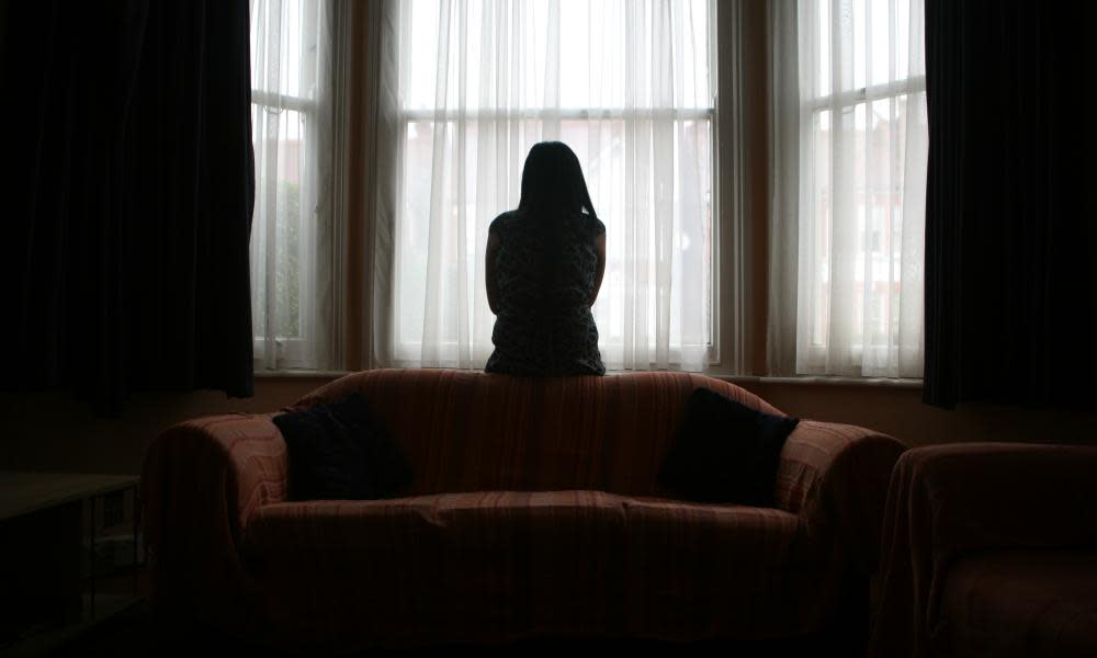 A young woman suffering from domestic violence stands alone in the bay window of her home