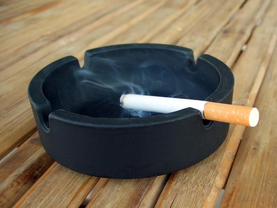 A black ashtray on a wooden table with a smoking cigarette on it.