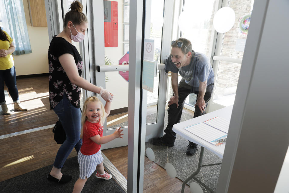 In this May 27, 2020 photo, Samantha Sulik, left, director of the Frederickson KinderCare daycare center, in Tacoma, Wash., looks on as Michael Canfield, right, waits in an entryway to pick up his daughter Aurora at the end of the day. As a precaution against the spread of the coronavirus, parents are not allowed to come into the building when they pick up their children. (AP Photo/Ted S. Warren)