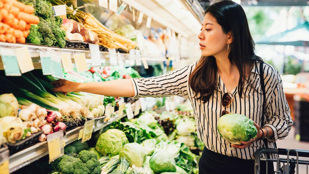 asian local woman buy vegetables and fruits in supermarket. young chinese lady holding green leaf vegetable and picking choosing green onion on cold open refrigerator. elegant female grocery shopping - Image