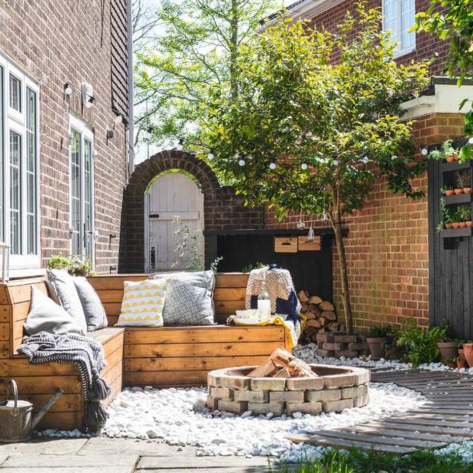 Small garden with tree, wooden outdoor seating, fire-pit and wooden boardwalk on white gravel, black wooden fence.