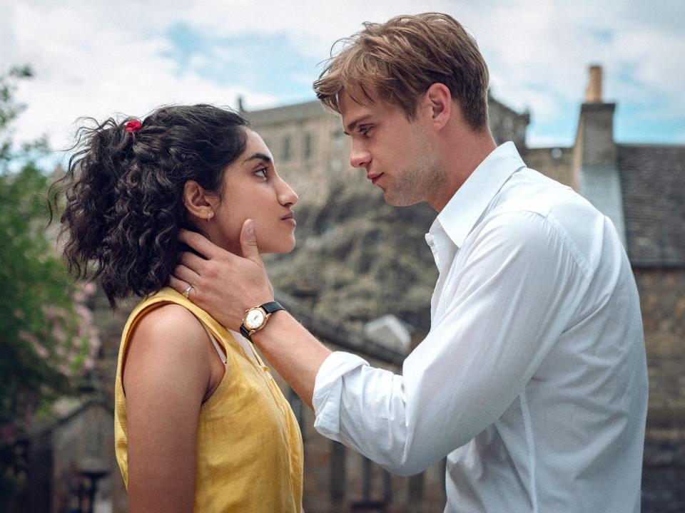 a young man cradles a woman's neck and jaw as they look intensely into each others' eyes face to face, standing outdoors in a picturesque village