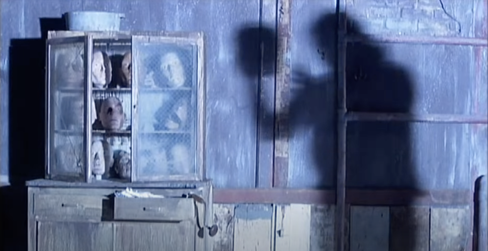 a shadow figure holding a mannequin head while other heads look from a bookcase