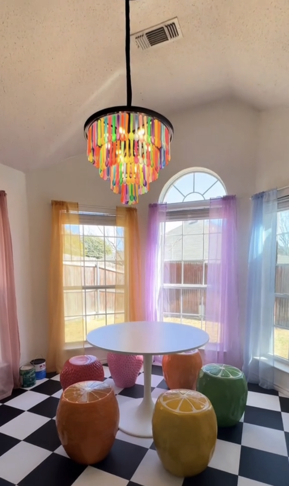 An overview of the breakfast nook: A chandelier made of colorful spoons above a white table with multicolored fruit chairs and in the background, curtains of different colors hang from windows