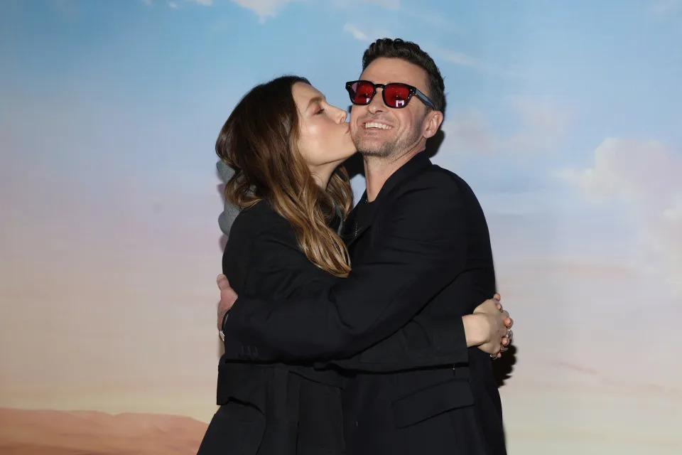 A woman wearing a black outfit kisses a smiling man's cheek while they embrace. The man is wearing a black outfit and red sunglasses
