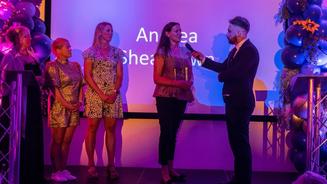 Andrea, a woman with long brown hair is interviewed on stage as she collects her award. Her name is on a purple screen behind her and she is flanked by two of her friends and the male host