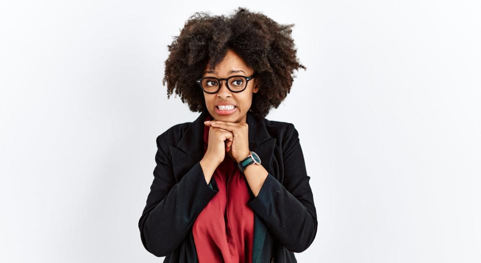 Woman with curly hair, glasses, biting fingernails in anxious pose, wearing a blazer, red top, and jeans