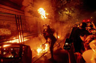 FILE - In this July 22, 2020, file photo, protesters throw flaming debris over a fence at the Mark O. Hatfield United States Courthouse in Portland, Ore. Until a year ago, the city was best known nationally for its ambrosial food scene, craft breweries and “Portlandia” hipsters. Now, months-long protests following the killing of George Floyd, a surge in deadly gun violence, and an increasingly visible homeless population have many questioning whether Oregon’s largest city can recover. (AP Photo/Noah Berger)