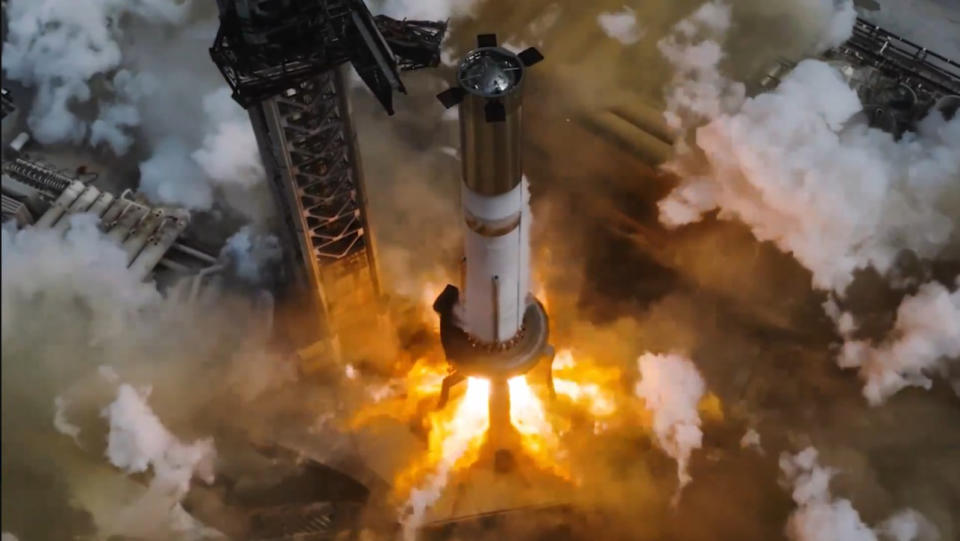  A tall chrome rocket booster stands next to a launch tower as huge explosions of fire burst from its engines below. Smoke billows in all directions. 