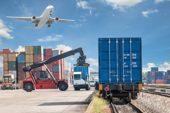 Shipping container being transferred from truck to freight train, with freight aircraft taking off in background.