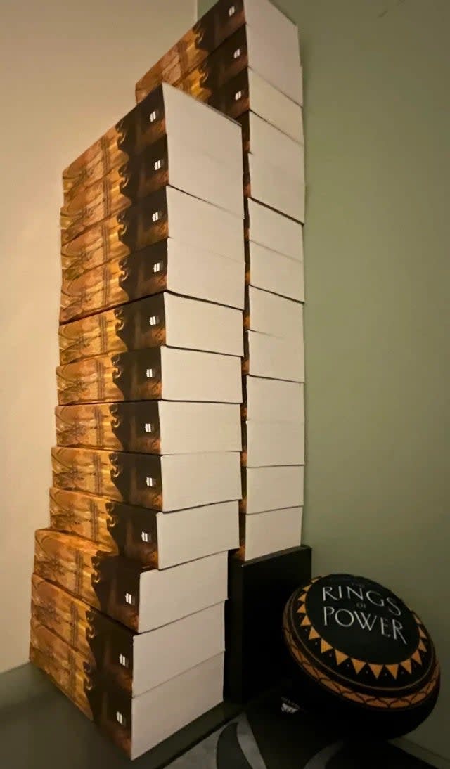 25 Lord of the Rings books are stacked on top of each other next to a Rings of Power pillow.