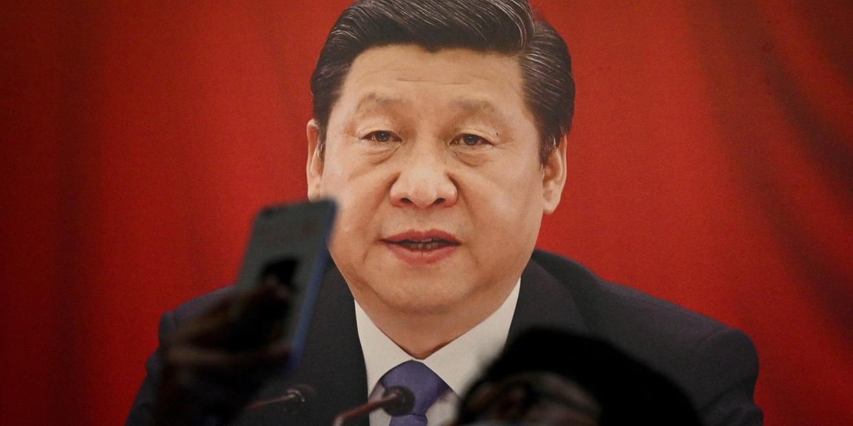 A man wearing a mask and holding a phone up in front of his face stands in front of a poster of Xi Jinping.