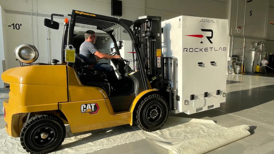  A yellow forklift carries a large white box with the words "rocket lab" on its side. 