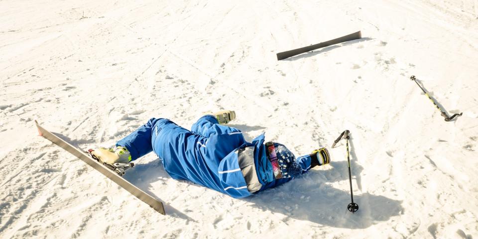 A skier wearing a blue ski suit, helmet, and goggles falls flat in the snow, with a ski and pole left behind.