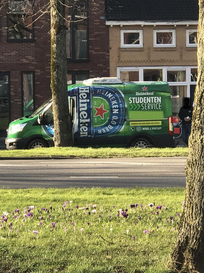 A Heineken-branded van with "Student Service" text, parked by a tree-lined street, near some flowering plants