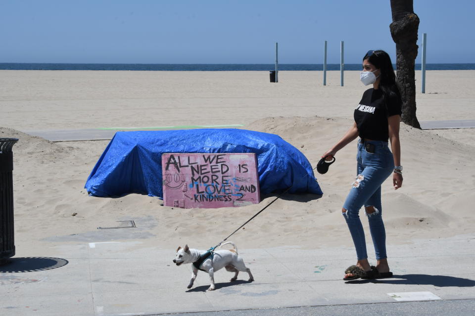 A lady wearing a mask due to the coronavirus pandemic   walks her dog past a sign that reads All we need is love along Ocean Front Walk at Venice Beach. Source: Getty